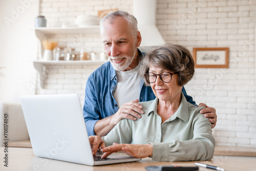 Active seniors concept. Caucasian old elderly senior wife and husband, couple spouses grandparents using laptop for e-banking, online shopping, remote work and telehealth at home kitchen