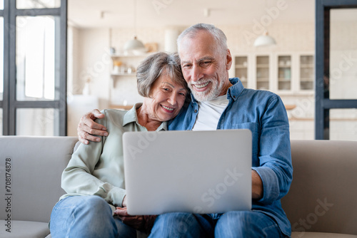 Caucasian senior happy old elderly grandparents couple spouses hugging embracing cuddling together while watching movie film series on laptop at home