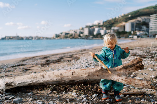 Little girl runs a stick along a snag lying on the seashore © Nadtochiy