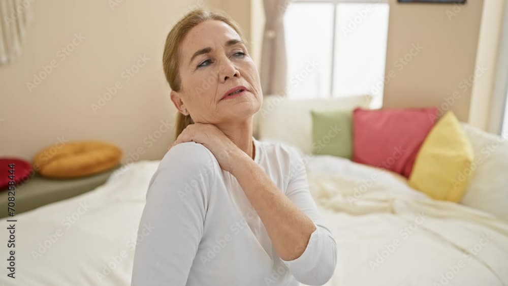 A mature woman experiencing shoulder pain in her cozy bedroom filled with colorful pillows. A mature woman experiencing shoulder pain in her cozy bedroom filled with colorful pillows.