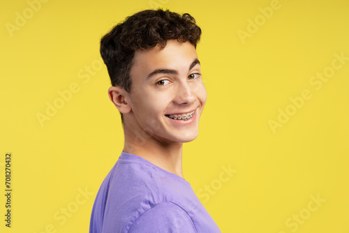 Portrait of smiling curly haired boy with dental braces looking at camera isolated on yellow background. Health care, orthodontic concept
