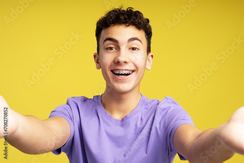 Portrait smiling boy, teenager wearing dental braces taking selfie isolated on yellow background. Happy young influencer recording video, communication online. Video call concept  photo