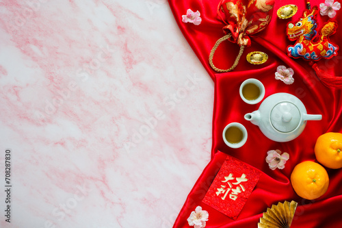 Tea set on red cloth background with red envelope packets or ang bao(word mean auspice), red bag, ingots(word mean wealth), hanging pendant dragon and oranges on marble table.