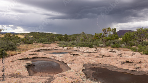 фотографія A distant lightning bolt drops from the heavy storm clouds over a mesa top in Utah's southwest desert while ephemeral pools on the sandstone surface wait for more rain to refill them