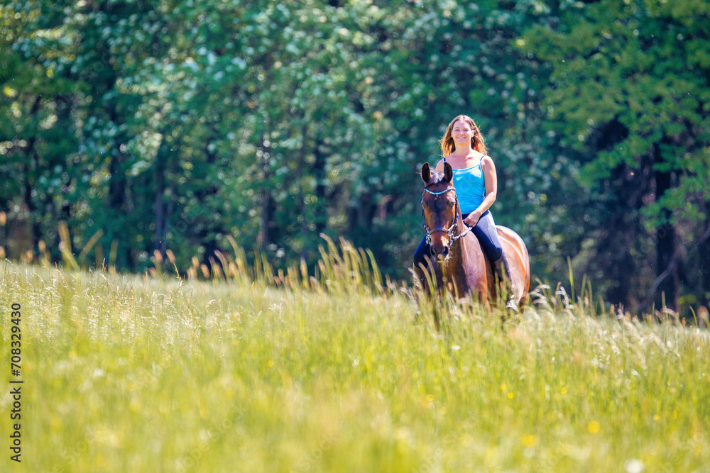 Young woman with brunette long hair rides bareback with her brown horse across a summer meadow, dressed in a blue tank top and riding pants with boots.