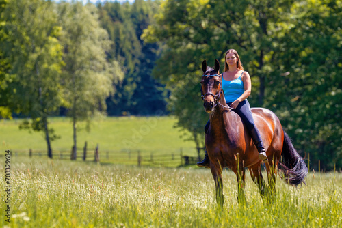 Young woman with brunette long hair rides bareback with her brown horse across a summer meadow, dressed in a blue tank top and riding pants with boots. © RD-Fotografie