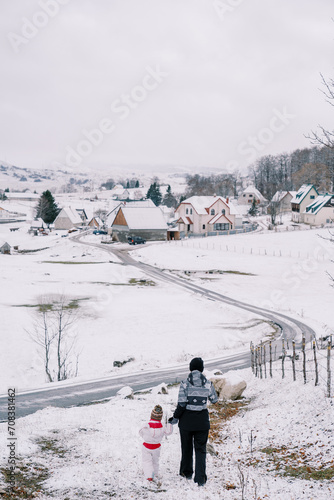 Mother and a little girl walk down a snowy hill holding hands towards a village road. Back view