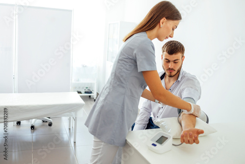 Nurse in medical uniform and transparent gloves checking blood pressure of young man in casual clothes. Brunette woman presses button on tonometer. Test tubes and folders with papers on background.