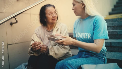 Caring volunteer donating hot food to homeless woman sitting on stairs, charity photo