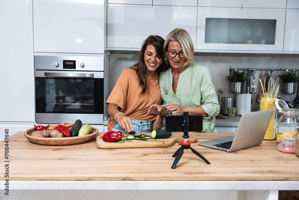 Obraz premium Grandma and daughter record a cooking vlog or podcast while chopping vegetables for a healthy vegetarian meal in the kitchen and stream it online. Vegan food preparation