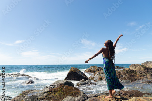 Beautiful woman, with braids and blue clothes, standing on the rocks at the beach posing for a photo