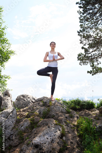 Attractive young woman doing a yoga pose for balance and stretching staying on top of high rock in the mountains