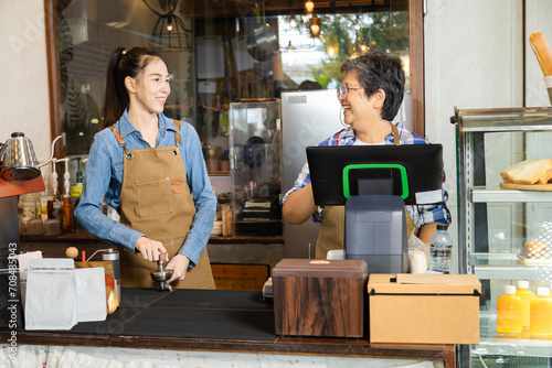 Young barista female hiring senior, elderly, 60s pensioner worker working in cafe bakery small business shop, two waitresses women standing behind coffee counter using technology cash register machine photo