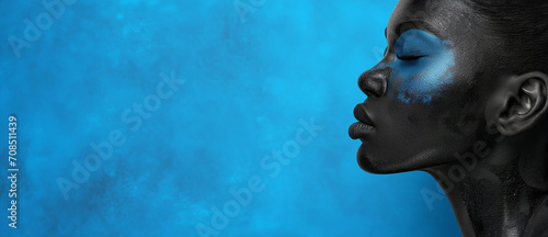 Beautiful African woman side profile painted with paints face view on bright blue wall studio background. Beauty, fashion, cosmetics concept