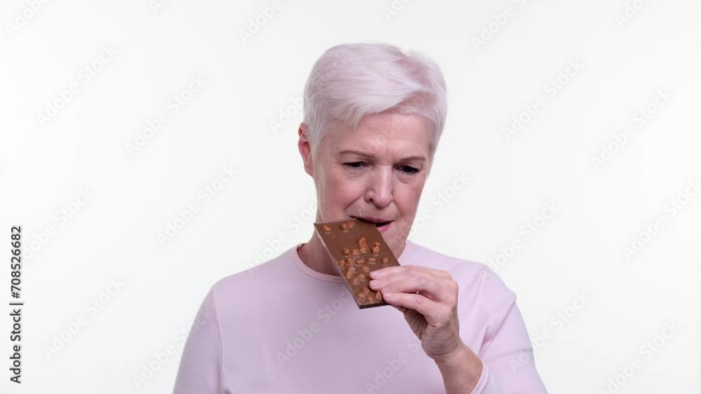 Senior woman takes a satisfying bite from a chocolate bar filled with crunchy nuts, eyes closing in sheer delight. The indulgent moment captures the joy of savoring a delectable treat. Senior woman takes a satisfying bite from a chocolate bar filled with crunchy nuts, eyes closing in sheer delight. The indulgent moment captures the joy of savoring a delectable treat.