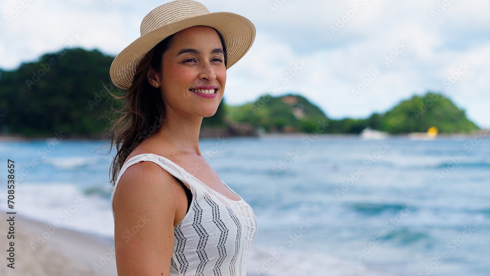 Beautiful girl with straw hat enjoying sunbath at beach. Close up face of young tanned woman with closed eyes enjoying breeze at seaside. Carefree latin woman smiling with ocean in background.
