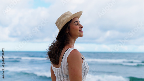 Beautiful girl with straw hat enjoying sunbath at beach. Close up face of young tanned woman with closed eyes enjoying breeze at seaside. Carefree latin woman smiling with ocean in background. 