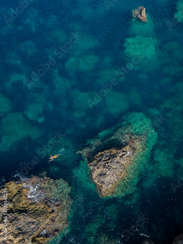 Single snorkeler in yellow swim trunks swimming in turquoise sea by the rocks in Malta aerial top down vertical
