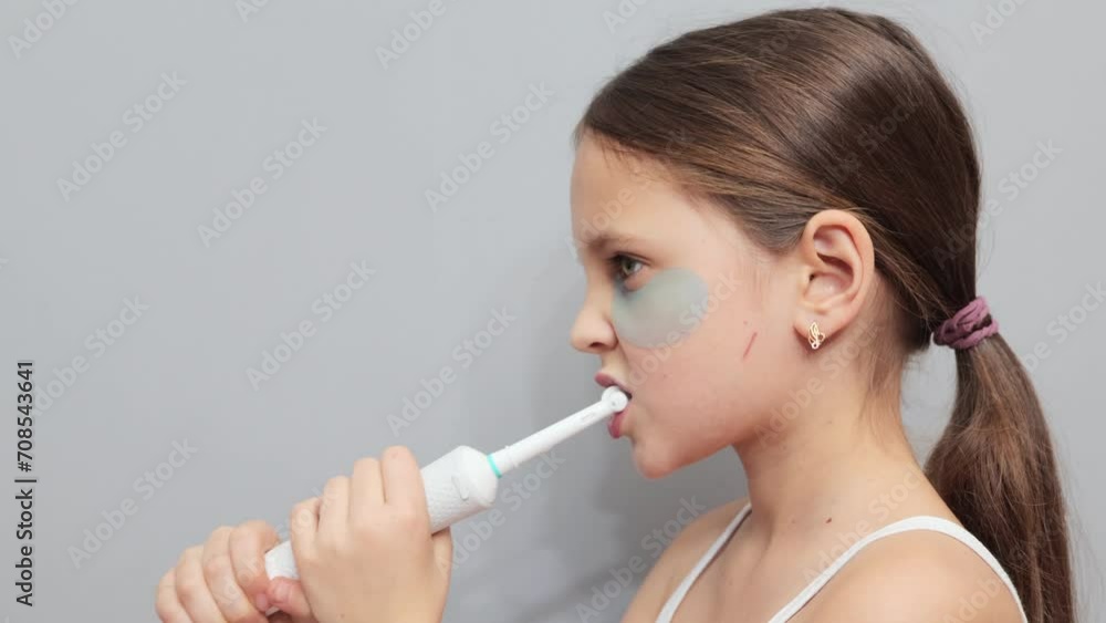 Side view of brown haired little girl with under eyes patches taking care of her oral health brushing her teeth with electric toothbrush isolated on gray background Side view of brown haired little girl with under eyes patches taking care of her oral health brushing her teeth with electric toothbrush isolated on gray background