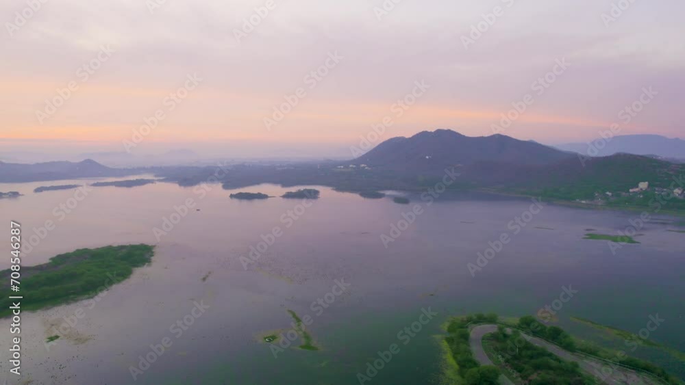 aerial drone shot at dawn dusk with road loop extending into fateh sagar lake with aravalli hills in distance hidden in fog showing cityscape of Udaipur Rajasthan aerial drone shot at dawn dusk with road loop extending into fateh sagar lake with aravalli hills in distance hidden in fog showing cityscape of Udaipur Rajasthan