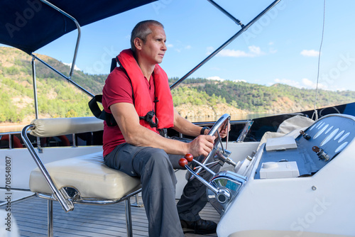 Young caucasian man steering motor yacht. Motor boat captain. Master of yacht. Yachting concept. Man wearing inflatable life jacket.