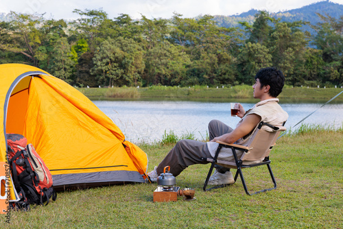 An Asian man sitting and drinking coffee near camping tent beside the lake and mountain in the sunshine morning with backpack and mobile solar cell