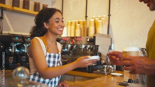 Woman wearing apron behind counter taking contactless card payment from customer in coffee shop or cafe - shot in real time