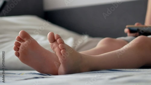 The bare feet of a child sitting in bed in the morning with a smartphone watching videos or playing games. Modern wireless technologies and the Internet.