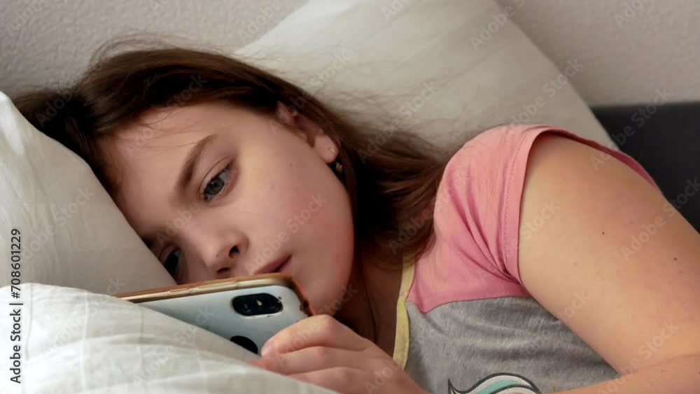 A little girl is sick, lying in bed weak and sad, looking into the frame in pajamas with disheveled hair, watching a video on a smartphone. Melancholy A little girl is sick, lying in bed weak and sad, looking into the frame in pajamas with disheveled hair, watching a video on a smartphone. Melancholy