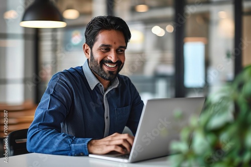 Happy Indian business man looking at laptop using computer working in office. Smiling busy professional businessman employee or company executive cheerful about online project goals, Generative AI