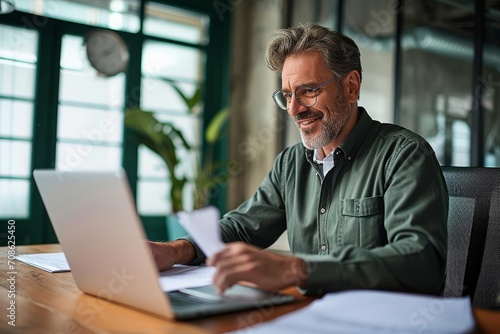 Smiling middle aged executive, mature male hr manager holding documents using laptop looking at pc computer in office at desk, checking financial data, Generative AI