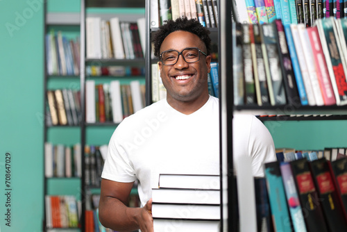 Portrait of a happy african male librarian carrying a stack of new books for readers. Dark-skinned guy in white t-shirt on the background of shelves with art books smiling cheerfully photo