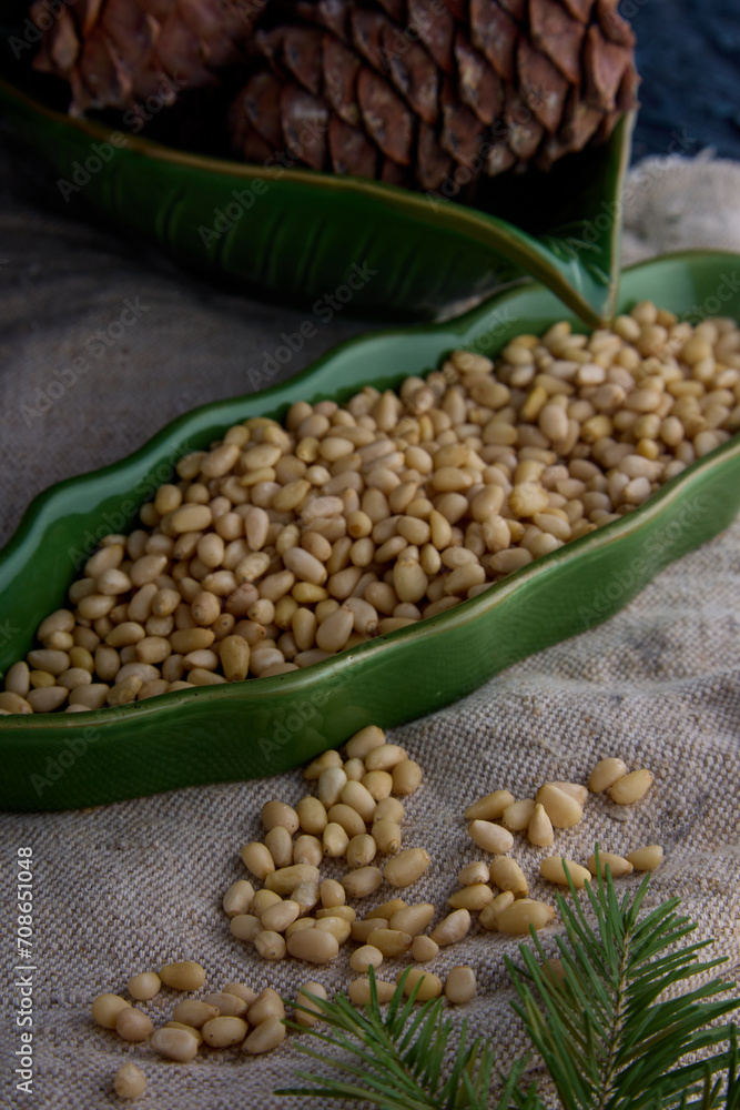 Obraz premium Tasty and healthy nuts rich in protein and oils close-up in green dishes. Peeled pine nuts and brown pine cone in green plates on old burlap. 