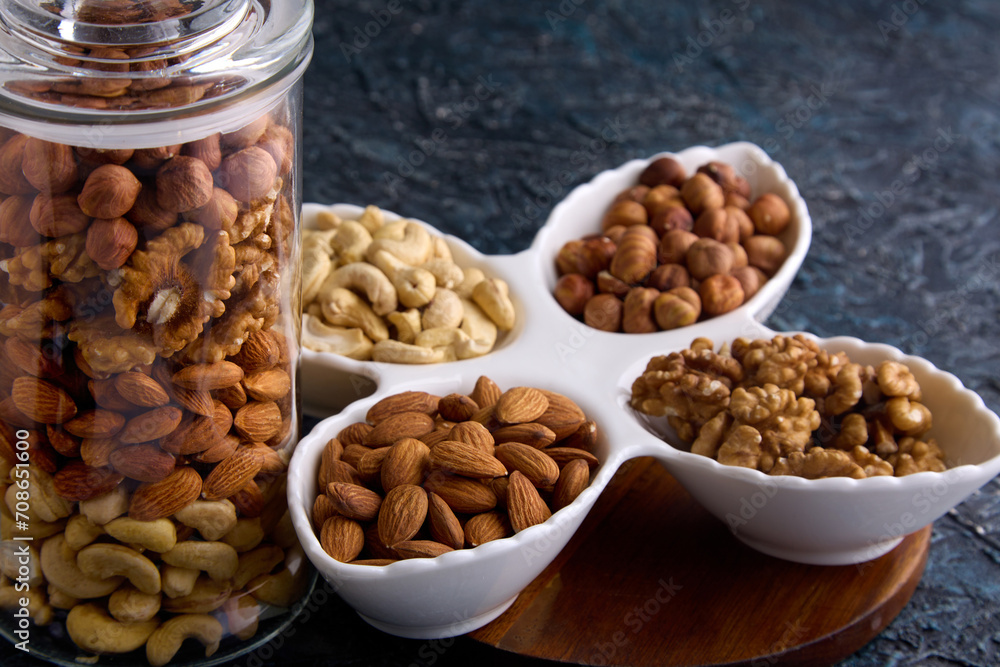 Glass transparent jar holds a mixture of different dried nuts next to a beautiful white plate on the table. Beautiful layout of nuts on a gray background in white dishes and a glass jar Glass transparent jar holds a mixture of different dried nuts next to a beautiful white plate on the table. Beautiful layout of nuts on a gray background in white dishes and a glass jar