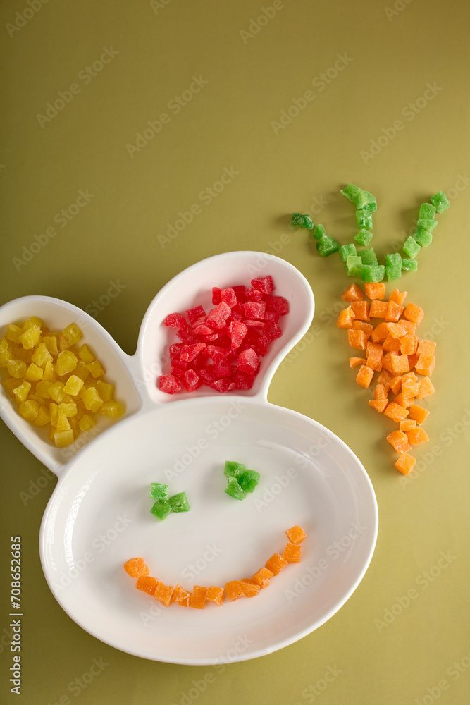 On children plate in shape hare's head made delicious colored candied fruits, smile and shape carrot laid out. Beautiful funny serving children's plate with delicious sweet sweets from candied fruits On children plate in shape hare's head made delicious colored candied fruits, smile and shape carrot laid out. Beautiful funny serving children's plate with delicious sweet sweets from candied fruits