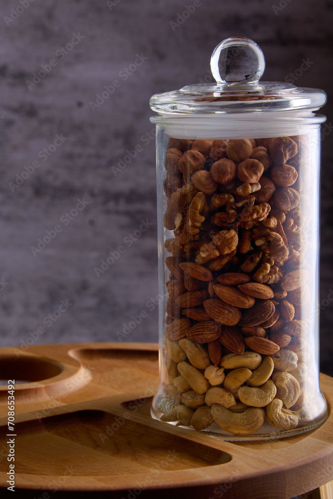 Walnuts, cashews, almonds, hazelnuts in a jar. On a wooden plate there is a transparent glass jar with a lid filled with assorted nuts. Storage of delicious and healthy nuts in a glass jar with a lid. Walnuts, cashews, almonds, hazelnuts in a jar. On a wooden plate there is a transparent glass jar with a lid filled with assorted nuts. Storage of delicious and healthy nuts in a glass jar with a lid.