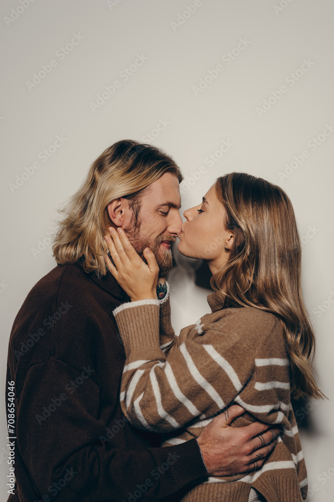 Happy young loving couple in cozy warm wear embracing and kissing while standing on beige background Happy young loving couple in cozy warm wear embracing and kissing while standing on beige background
