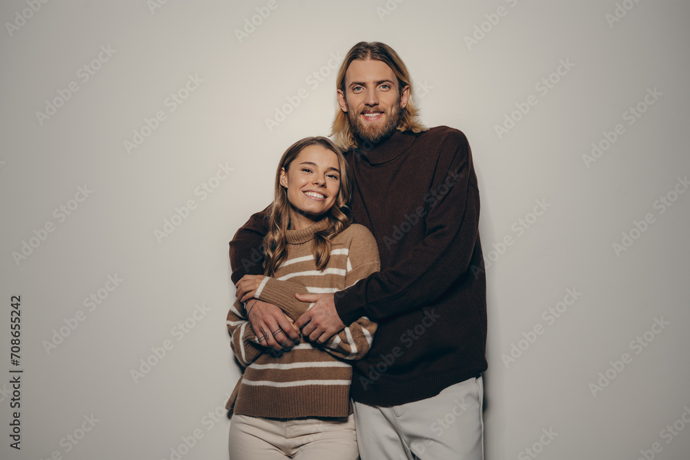 Fototapeta premium Fashionable young loving couple embracing and looking at camera with smiles on beige background