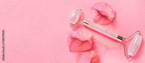 Face roller over pink background with flowers. Jade roller facial lifting massage tool, anti aging treatment, gua sha pink quartz rolling device for skin care, self massaging accessory. Beauty concept photo