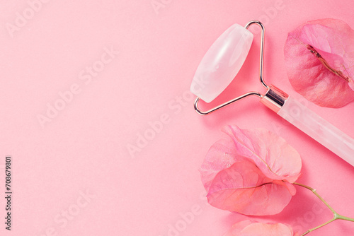 Face Jade roller over pink background with flowers. Facial lifting massage tool, anti aging treatment, gua sha pink quartz rolling device for skin care, self massaging accessory. Beauty concept photo