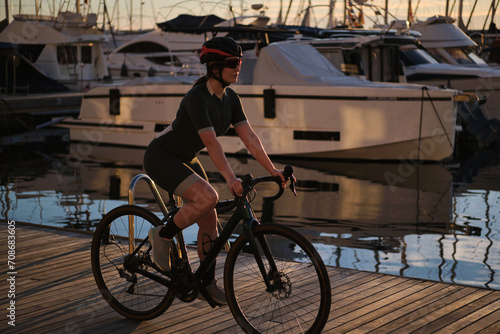 Woman cyclist wearing cycling kit and helmet ,riding a bicycle against the backdrop of a yacht club during the sunset. Cycling training in evening time. Sport motivation. Active lifestyle concept.