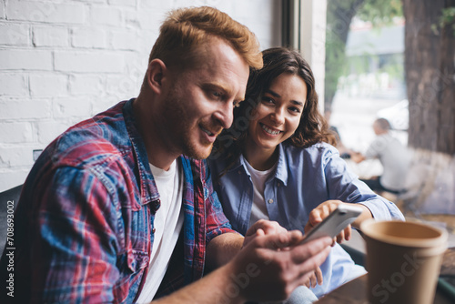 Portrait of cheerful Caucasian blogger smiling at camera while bearded boyfriend searching website on modern smartphone technology, happy couple in love using cellphone device for social networking photo