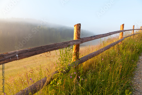 Wooden fence near the meadow on a foggy morning, sun shines through the fog. Ukraine, Carpathians.