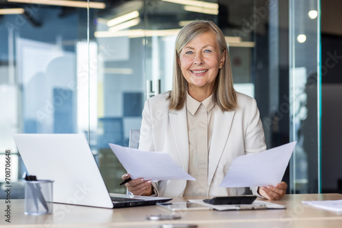 Confident senior businesswoman with documents at office desk  smiling at camera  professional workplace.