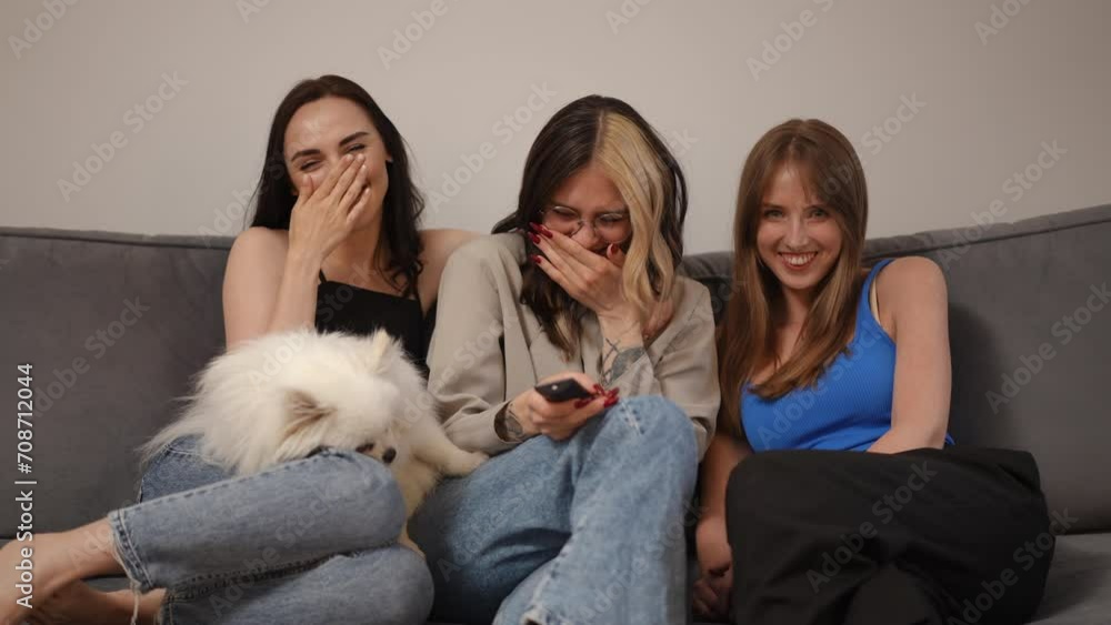 Three caucasian girlfriends are sitting on the sofa and burst into laughter, looking at the camera. A white Spitz sleeps on the lap of one of the girls. A girl with glasses holding a TV remote control Three caucasian girlfriends are sitting on the sofa and burst into laughter, looking at the camera. A white Spitz sleeps on the lap of one of the girls. A girl with glasses holding a TV remote control