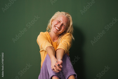 Happy satisfied young woman in casual outfit smiling at camera sitting on the ground in a studio against a green background. © Dorde