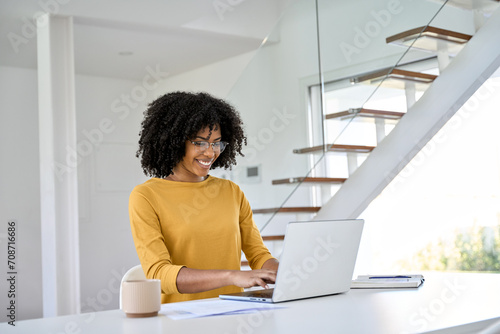 Smiling young pretty happy African American woman wearing glasses using laptop computer sitting at home table hybrid working online, female student elearning, browsing typing at pc. photo