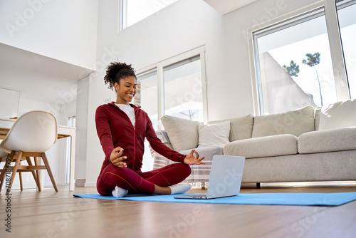Young happy healthy African American woman wearing sportswear sitting on floor at home doing yoga breathing exercise, meditating learning online training fitness online class on computer. photo