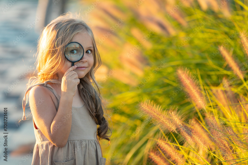 Cute adorable Caucasian girl looking at plants grass in park through magnifying glass. Child looking through a magnifying glass on the grass in summer day. Child education, early development. concept.
