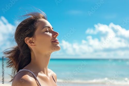 Beautiful young woman raises her head towards the sun to enjoy the heat in summer, with the beach and blue sky in the background.